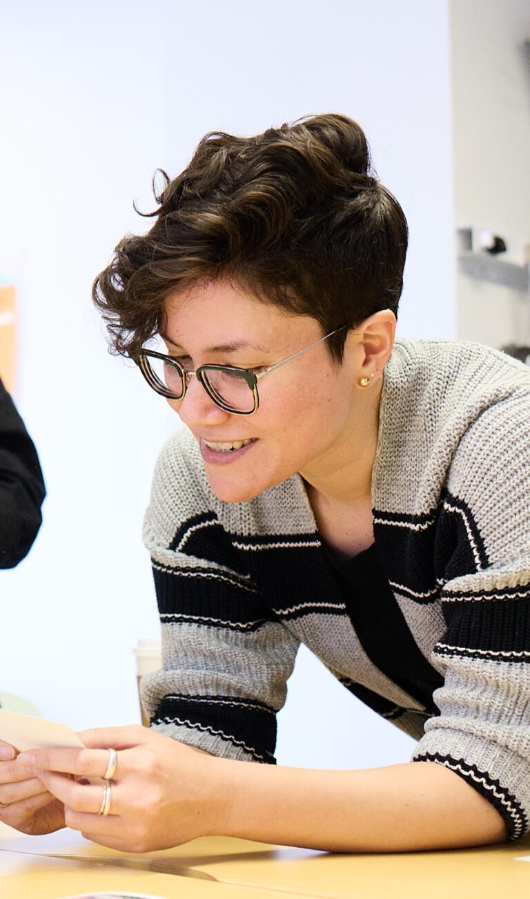 Photograph of Alex Kostiw. A light-skinned person with black-and-gold-rimmed glasses and short brown hair contemplates a piece of paper.