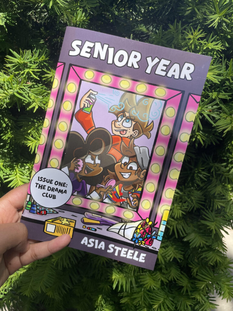 Photograph of the cover of Senior Year Issue One: The Drama Club by Asia Steele. Three characters prepare themselves at a lighted vanity table; a bouquet of flowers and various cosmetics are scattered on the table.