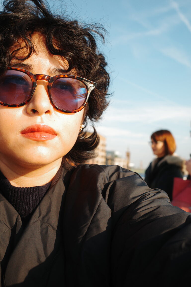 Photograph of Elle Shivers. A person with short, wavy dark hair and sunglasses gazes into the distance, framed by a blue sky.